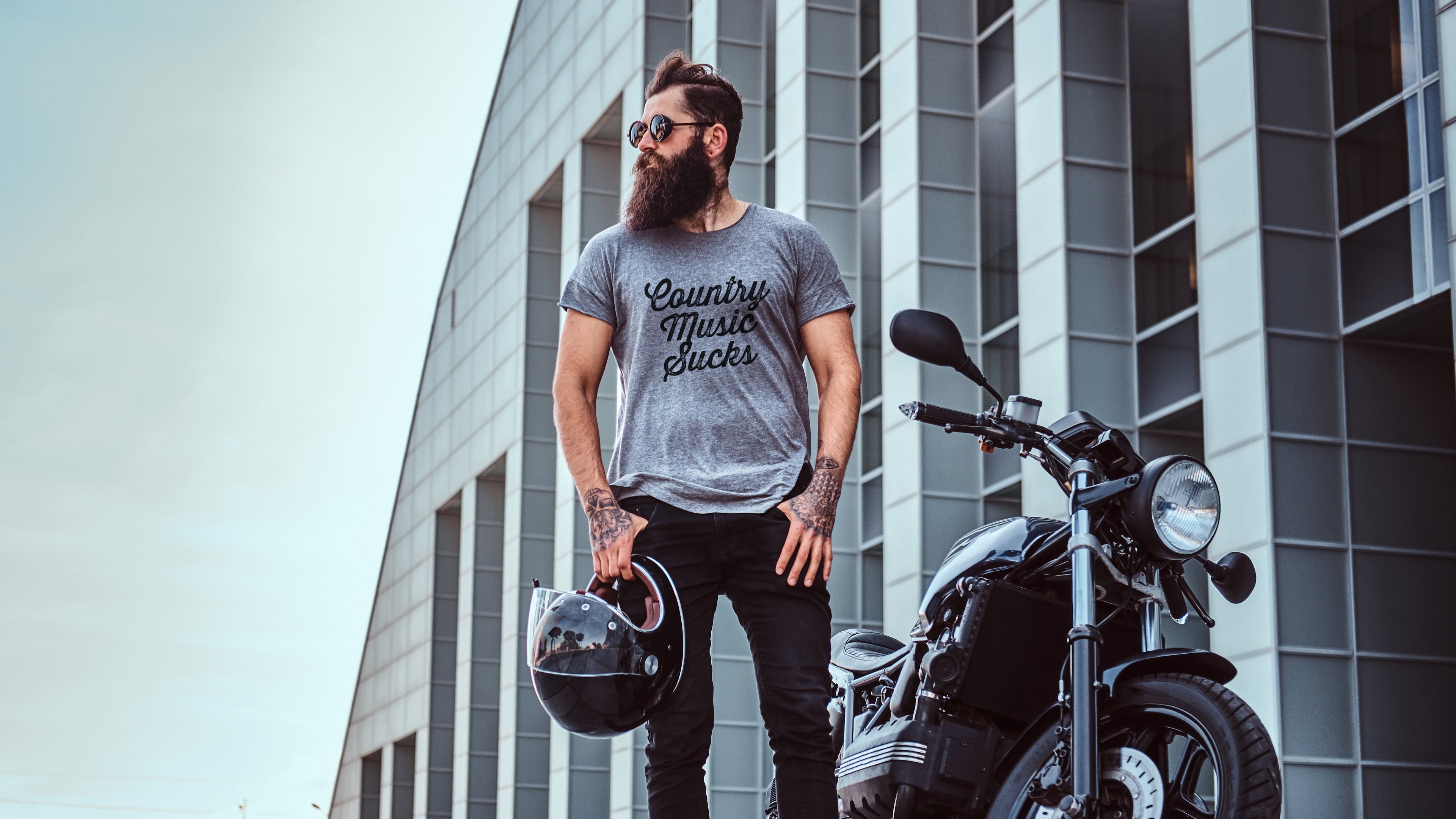 Man with a beard standing next to a motorcycle wearing a t-shirt with text, holding a helmet.